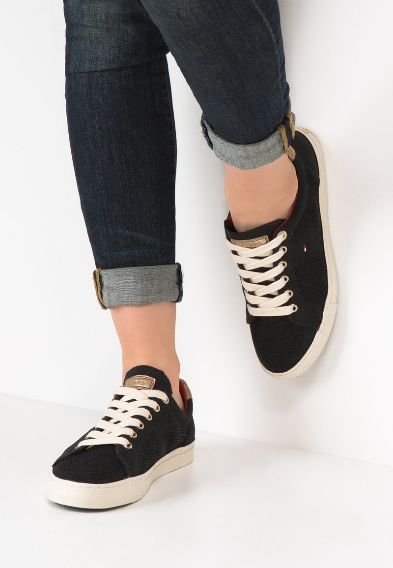 Sneakers a rete nera con lacci crema e suola in gomma bianca. Presentano dettagli dorati e una superficie testurizzata. I jeans sono arrotolati ai polsi.