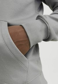 Grauer Sweatshirt mit geripptem Bündchen und Kängurutasche. Der Stoff ist glatt mit einer leichten Textur, die die Nahtdetails der Tasche hervorhebt.
