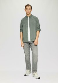 Groene button-up shirt over een wit T-shirt, gecombineerd met grijze jeans en lichtgekleurde sneakers. Model staat tegen een neutrale achtergrond.