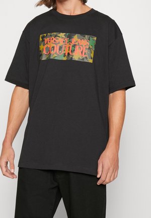 Osoba nosząca czarną koszulkę z krótkim rękawem z prostokątnym logo w kamuflaż i wzór gwiazd z napisem "Versace Jeans Couture" czerwonymi literami.