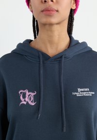 Mørkeblå hettegenser med rosa grafisk tekst og logo på brystet. Har justerbare snorer og en avslappet passform.