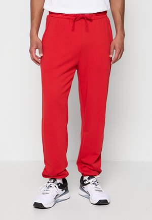 Man die felrode joggingbroek en witte sportschoenen draagt, staand tegen een effen achtergrond.