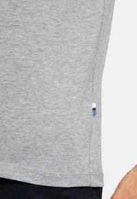 Maglietta grigia con una texture morbida, dotata di una corta fessura laterale e un'etichetta decorativa blu navy, bianca e blu sull'orlo.