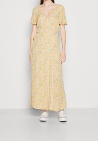 Robe portefeuille florale jaune à manches courtes, avec un décolleté en V, un tissu fluide et une silhouette longue et à volants. Portée avec des baskets blanches.
