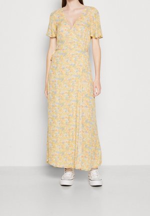 Geel bloemen wikkeljurk met korte mouwen, voorzien van een v-hals, vloeiend materiaal en een lange, gelaagde silhouet. G dragen met witte sneakers.