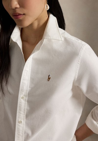 Femme portant une chemise blanche boutonnée avec un petit logo brodé de joueur de polo sur la poitrine et une boucle d'oreille en coquillage blanc à une oreille.