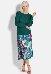 Grüner gestrickter Pullover mit strukturiertem Muster, kombiniert mit einem blumigen Midi-Rock, der pinke und türkisfarbene Blumen aufweist, und pinken Riemchensandalen.