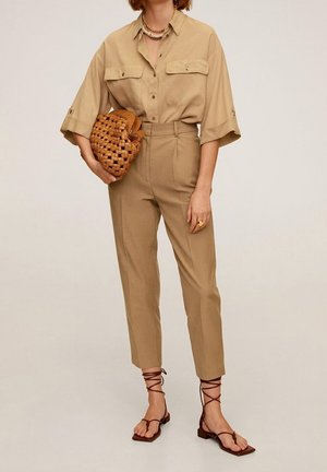 Femme portant une chemise beige boutonnée et un pantalon taille haute, tenant une pochette marron tressée avec des sandales lacées et des colliers superposés.