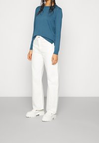 Blauer Strickpullover, weiße Jeans mit geradem Bein und weiße Slipper mit dicken Sohlen. Flache Stofftextur, lässige Silhouette.