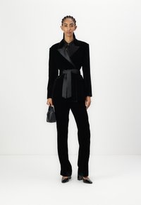 Black velvet blazer with satin lapels, paired with tailored black pants. Model holds a small black handbag, wearing sleek, pointed heels.
