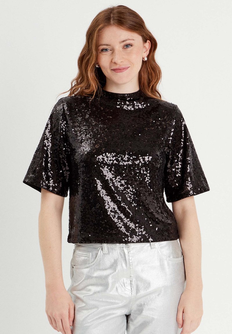 Top corto de lentejuelas negras con un cuello alto y mangas cortas, que presenta una textura brillante y lentejuelas que reflejan la luz, combinado con pantalones plateados.