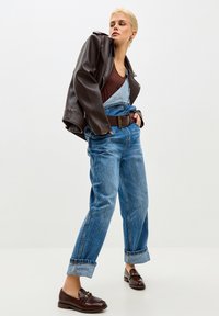 Žena odjevena u plavu traper kombinezon s jednom naramenicom otkopčanom, smeđom kožnom jaknom prebačenom preko ramena i smeđim loafericama, pozira samouvjereno.