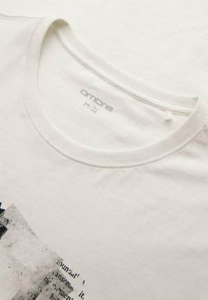 Camiseta blanca de cuello redondo con etiqueta de la marca "ombre" y diseño gráfico parcial con texto en negro y gris en la parte delantera.