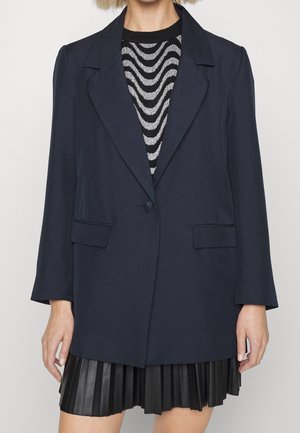 Frau trägt ein marineblaues Blazer mit einem Knopf über einem schwarz-weißen, wellenförmig gemusterten Oberteil, kombiniert mit einem schwarzen Faltenrock.