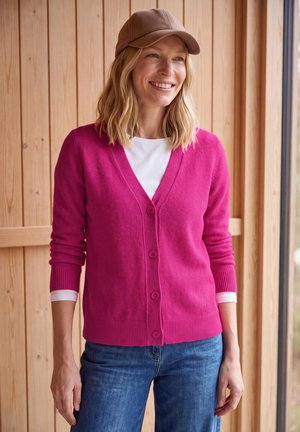Mujer sonriente en el interior con una gorra marrón, cárdigan rosa brillante abotonado, camisa blanca de manga larga y vaqueros azules, de pie junto a una pared y ventana de madera.