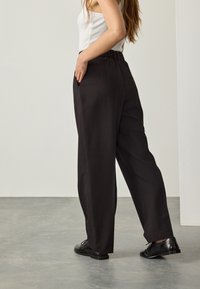 Pantalon noir ample avec une ceinture élastique, des poches sur les côtés et une texture de tissu lisse, associé à des chaussures noires.