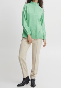 Pull-over en maille vert menthe avec col montant et demi-fermeture éclair, associé à un pantalon beige sur mesure et des bottines noires. Texture lisse.