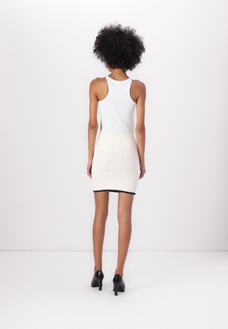 Biała bluzka na ramiączkach zestawiona z teksturowaną, kremową spódniczką mini z czarnym wykończeniem. Modelka stoi w czarnych szpilkach na jednolitym tle.