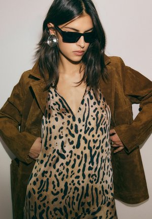 Žena nosi čierne slnečné okuliare, veľké strieborné náušnice, šaty s leopardím vzorom a hnedú semišovú bundu s rukami v kapse.