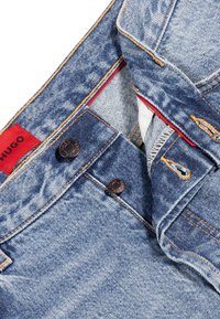 Pantalones vaqueros de mezclilla azul con una textura descolorida, que presentan botones de cobre y costuras naranjas. La etiqueta de la marca es roja con texto blanco.
