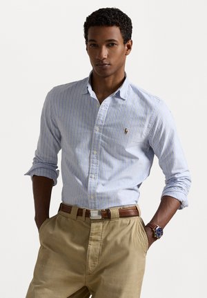 Világoskék és fehér csíkos, gombos ing klasszikus gallérral, felgöngyölt ujjakkal és egy kis logóval a mellkason, khaki nadrággal párosítva.