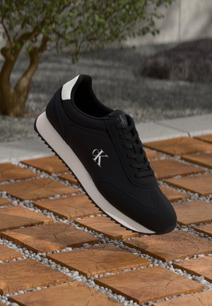 Basket Calvin Klein noire avec logo et semelle blanche, flottant au-dessus de carreaux carrés en bois avec de petits cailloux blancs entre eux en extérieur.