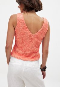 Coral sleeveless top with a V-back, textured with rose patterns, soft fabric, and delicate frayed edges. Paired with white linen pants.