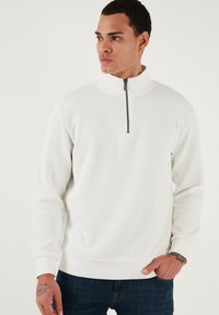 Biely rebrovaný sveter s vysokým golierom a polovičným zipsom. Obsahuje dlhé rukávy a voľný strih, skombinovaný s tmavými džínami.