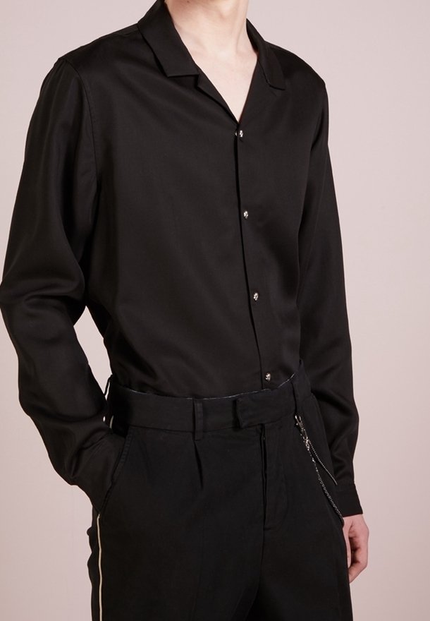 Homme portant une chemise noire à manches longues boutonnée, rentrée dans un pantalon noir avec un accessoire chaîne sur le côté, debout devant un fond uni.