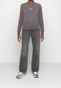 Felpa grigia in cotone con collo rotondo e logo ricamato sul petto, abbinata a jeans a gamba larga grigio scuro e sneakers con motivo.