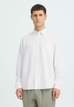 Junger Mann trägt ein weißes, langärmliges Hemd mit Knöpfen und Brusttasche sowie hellgrüne Hose, stehend vor einem schlichten weißen Hintergrund.