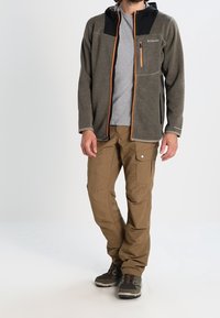 Man wearing gray Columbia fleece jacket with orange zipper, gray shirt, tan cargo pants, and gray hiking shoes standing against white background.