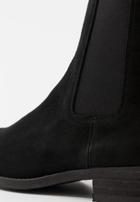 Svart mockaboot med ankellängd som har elastiska sidopaneler, slät yta, en låg klack och detaljerad söm längs kanten.