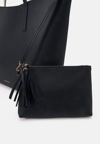 Black faux leather tote bag with textured surface, featuring long handles and a small matching pouch with a tassel accent. Gold hardware details.