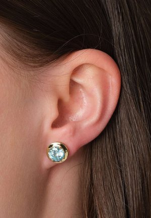 Goldfarbene Steckerohrringe mit einem großen, hellblauen Edelstein, der in einer glatten runden Fassung sitzt und nahe am Ohr positioniert ist.