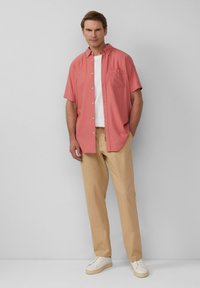 Korte mouwen koraalkleurige overhemd met een zak, gecombineerd met beige broek en witte sneakers, tegen een effen grijze achtergrond.