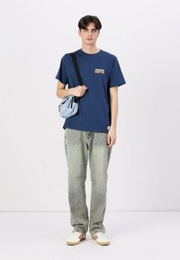 Marineblå t-shirt med et lille guldgrafik, lyseblå crossover taske, slidte grå jeans og hvide sneakers med røde detaljer.