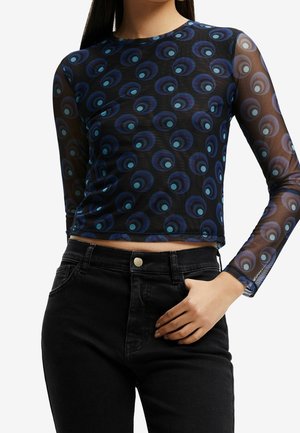 Langærmet mesh bluse med rund halsudskæring, prydet med et blåt og teal cirkulært mønster på en sort baggrund. Parret med sorte højtaljede jeans.