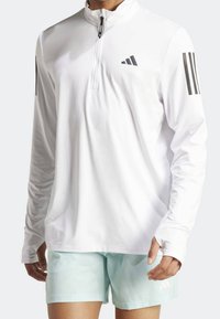 Biała długorękawowa koszulka sportowa z ćwierć zamkiem, z czarnymi trzema paskami na rękawach oraz logo marki na piersi.