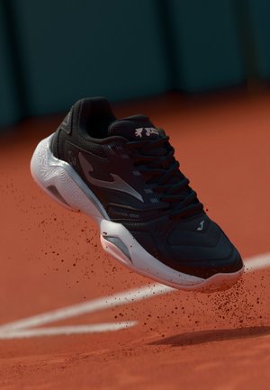 Scarpa sportiva nera con dettagli grigi, parte superiore testurizzata, suola bianca e dettaglio del logo. Posizionata sopra una superficie rossa con particelle di polvere.