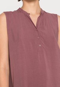 Blusa senza maniche in un tenue colore malva con una patta e tre bottoni. Tessuto morbido, leggero splendore e dettagli del colletto piegato sono visibili.