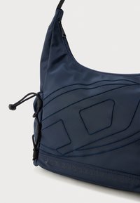 Sötétkék nejlon válltáska texturált felülettel. Díszítő varrással, felső cipzárral és állítható zsinórzáras oldalsó záródással rendelkezik.