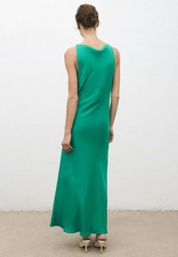 Vrouw met een bun, gekleed in een mouwloze groene maxi-jurk en beige hakken, staand tegen een effen witte muur.