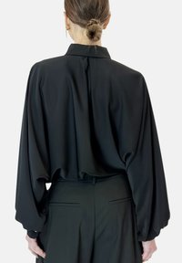 Blusa nera con maniche ampie e fluide e colletto alto. Realizzata in tessuto morbido, presenta un retro leggermente plissettato. Abbinata a pantaloni sartoriali.