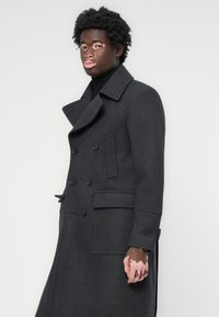 Manteau en laine gris foncé à double boutonnage avec grands revers, deux poches avant et accents de boutons. Présente une coupe ajustée et une texture lisse.