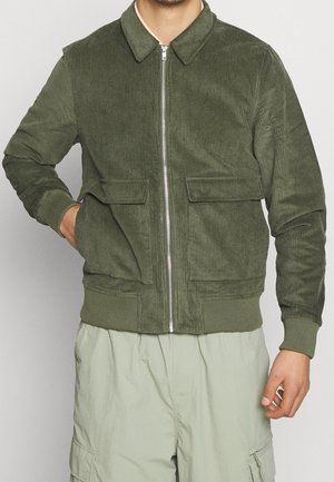 Uomo che indossa una giacca in velluto a coste verde oliva con zip e tasche a pattina e pantaloni cargo verde chiaro su sfondo semplice.