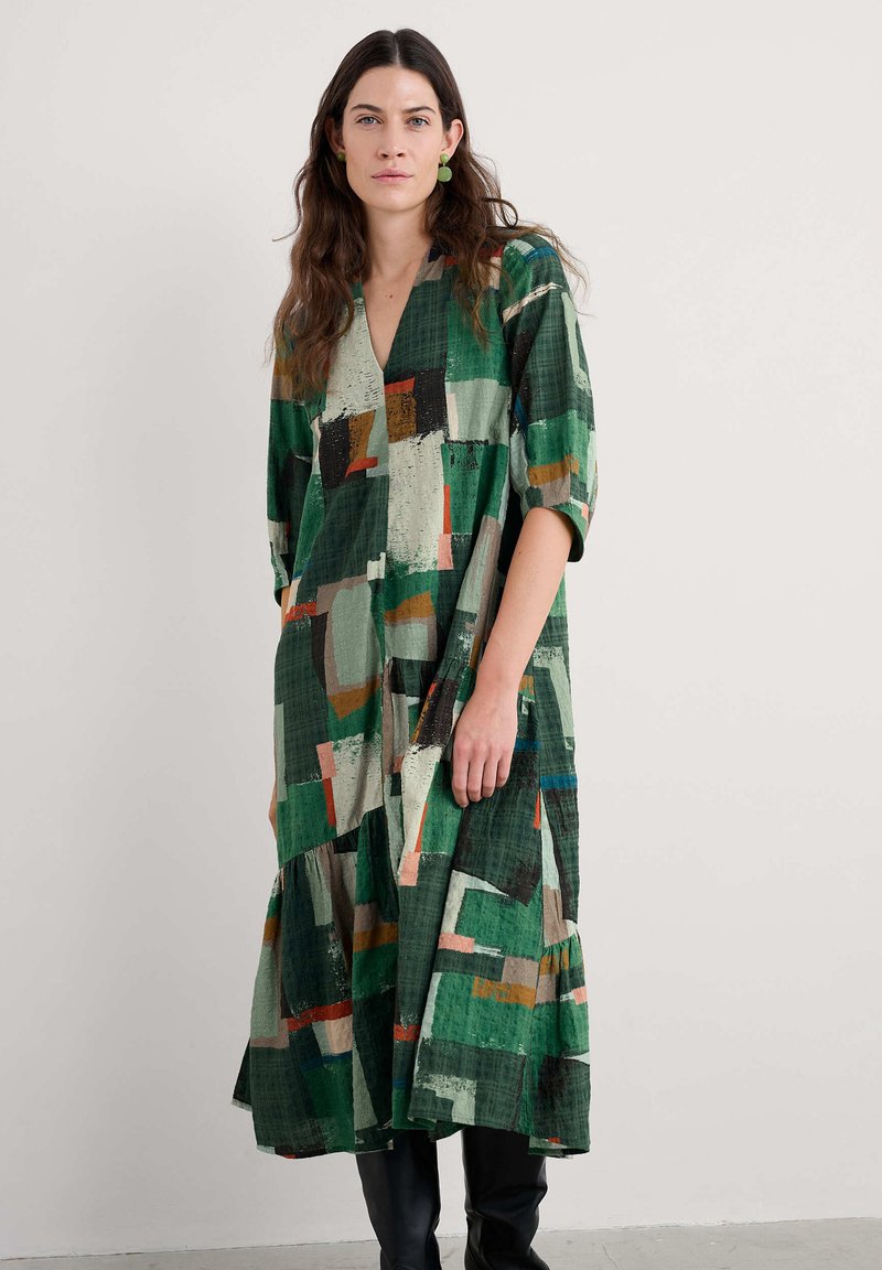 Mujer vistiendo un vestido midi de patrón abstracto en verde con mangas hasta el codo y botas negras hasta la rodilla, de pie contra un fondo liso.