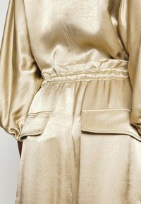 Conjunto dorado satinado con cinturilla elástica, mangas abullonadas y bolsillos con solapa en los pantalones, mostrado desde atrás a la altura de la cintura.