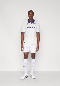 Biely futbalový dres s modrým golierom a potlačeným textom, doplnený o biele šortky a ponožky, s čiernym logom Nike a futbalovými kopačkami.