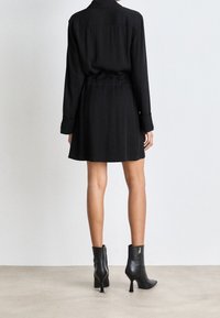 Donna in piedi di spalle di fronte a un muro, con un vestito lungo nero a maniche lunghe e stivaletti neri con tacco su un pavimento chiaro.